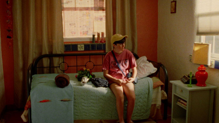 Nicole sits on a bed in a sunlit bedroom in Marlene Emilia Rios’ short film Lucia & Nicole.