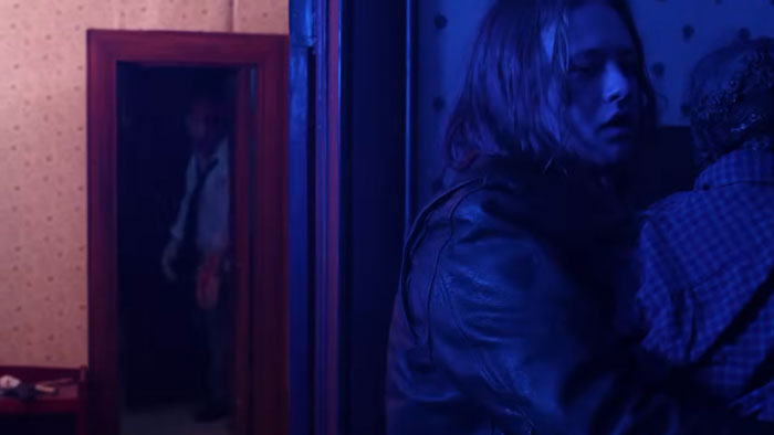 Danny (Sam Christensen) holds The Corpse (Sam Christensen) in a blue-lit hallway as a figure appears in a mirror.