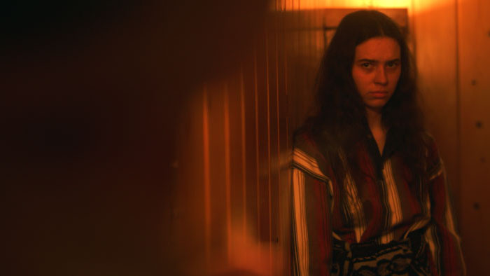 Freya stands in a dim orange-lit hallway in The Girl Inside The Photograph (2026). 