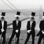The Residents wearing eyeball head masks and tuxedos seated in front of the Golden Gate Bridge in San Francisco