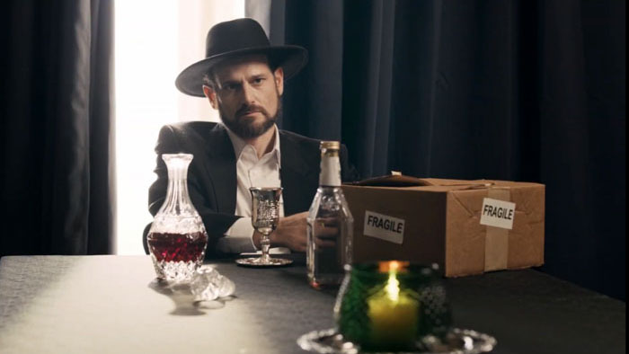 A man in a wide-brim hat sits at a table with a decanter, bottles, a candle, and a cardboard box labeled “FRAGILE” in Tooth Shop Fiasco (2026).