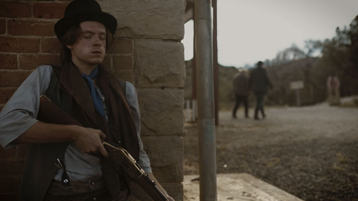 Francisco Zurcher as Billy the Kid leans against a brick wall holding a rifle in Blackwater Draw: The Making of Billy the Kid.