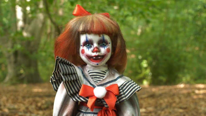 A sinister porcelain clown doll with red hair and white face paint stands in a forest in Lost Dolls 2.0.