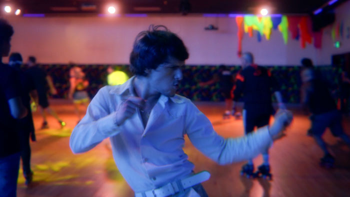 A young man in a white shirt dances at a neon-lit roller rink in The Floor Remembers.