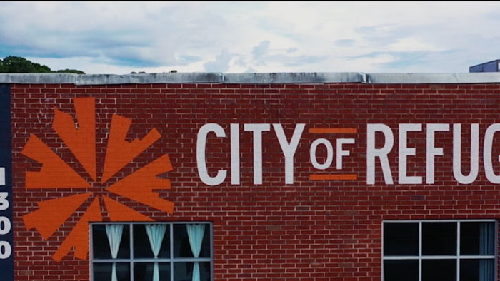The City of Refuge building with its orange logo painted on a red brick wall in The Greatest of These.