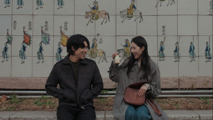 A man and woman laugh together on a street bench in front of a tiled mural in Before the Call.