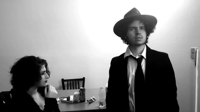 Adrian Chromstone (Bradley Rabinowitz) stands over Nikki Donovan seated at a table in RA Noir.