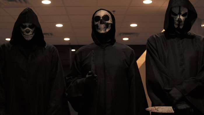 Three masked freaks in black robes threaten victims in We Can’t Leave.