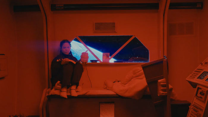Jeliza Zapanta in a blue spacesuit and headphones looks down inside the escape pod in A Light In The Ether.