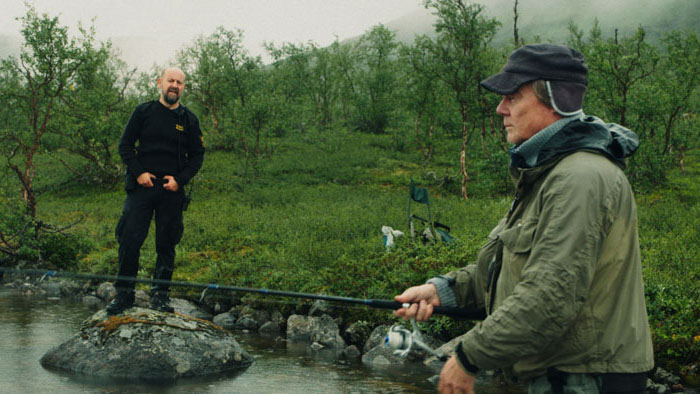 The customs officer watches as the elderly fisherman casts a line across a rocky stream in Borderline (På Grensa).