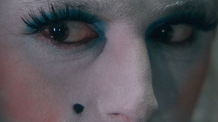 A close-up of a mysterious character with dramatic teal eye makeup and a beauty mark in Breakfast at Berghain.