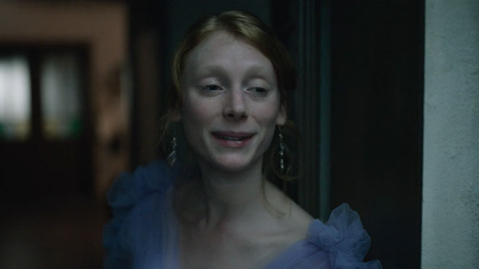 Nore (Luna Jordan) smiles through glass wearing a blue ruffled dress in Easy Girl.