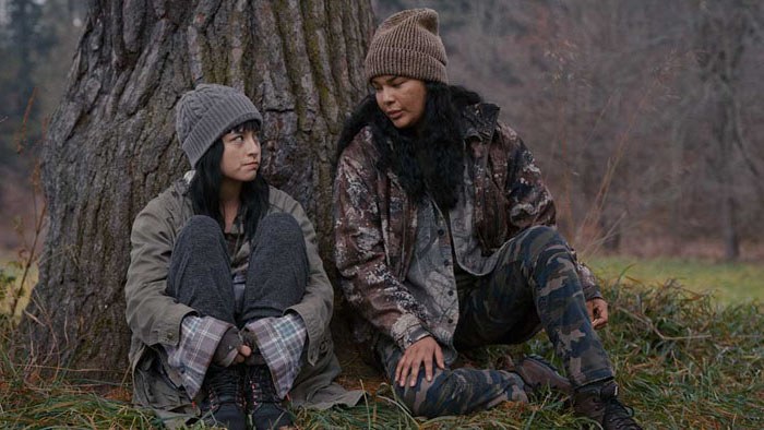 Nika (Ellyn Jade) and Madison (Star Slade) sit beneath a tree in the woods in Nika & Madison. 