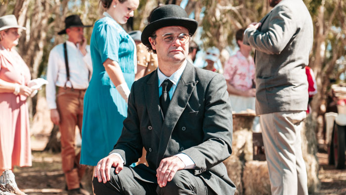The Bank Manager played by Gyton Grantley sits in a dark suit at an outdoor gathering in The Bank Manager.