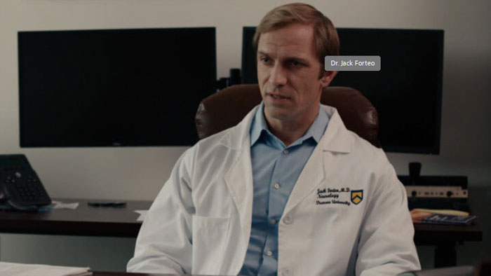 Dr. Jack Forteo (Andy Evans) sits at his university office desk in a lab coat in The Goose That Laid the Golden Egg.