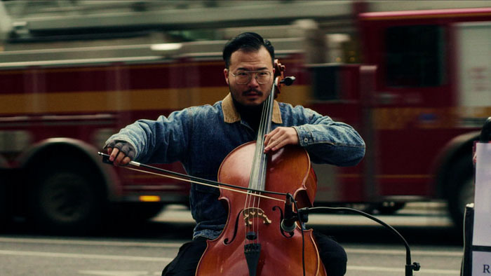 Kevin Zi-Xiao He plays cello on the street with a blurred fire truck passing behind him in To Busk or Not to Busk.