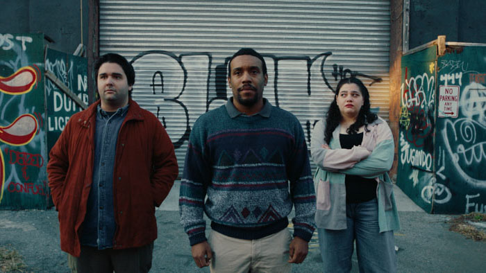 Zach (Elijah Lawrence) stands with two roommates in front of a graffiti-covered wall in Hamilton House.
