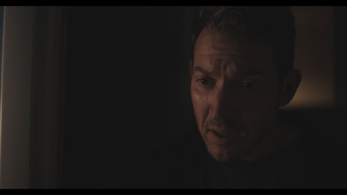 Alex (Matt Bailey) looks troubled in a dimly lit room in Open A Eye.