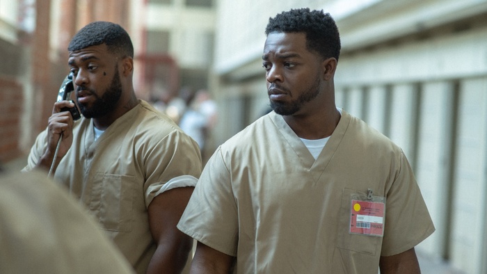Ricky walks through a prison corridor beside another incarcerated man using a telephone. 