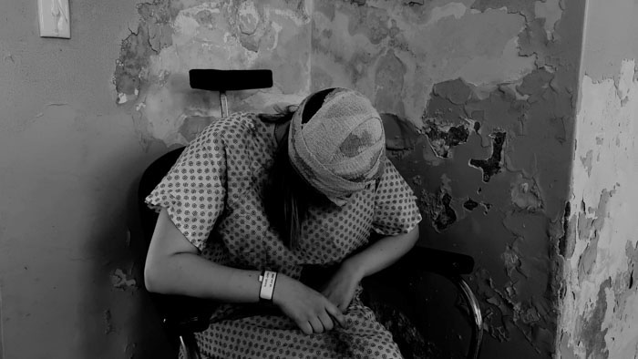A bandaged patient slumps in a wheelchair inside a decaying medical facility in The Cerebral Organoid.