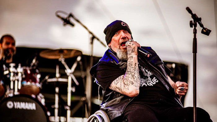 Paul Di'Anno sings into a microphone while performing from a wheelchair onstage.
