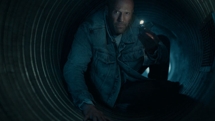 Cole Reed (Jason Statham) crawls through a dark ventilation shaft holding a lit lighter in Mutiny.