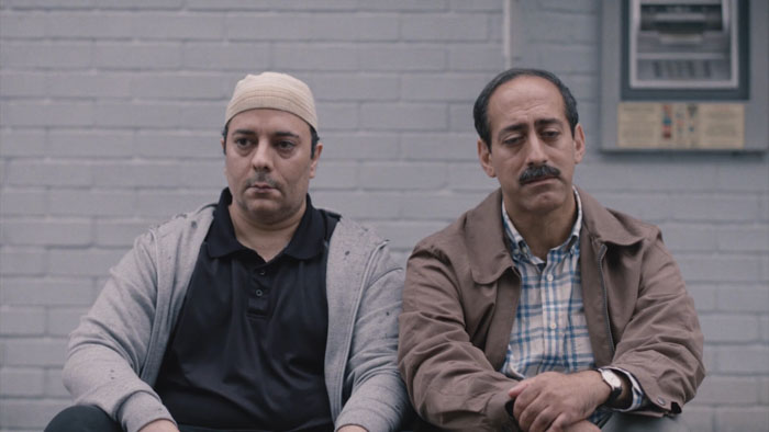 Shamsul (James Rana) sits with another man against a brick wall in Strays.