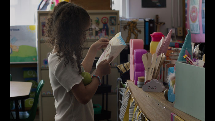 Ava (Soleila Shea) reaches for a drawing on a classroom shelf in Tears of Knowing.