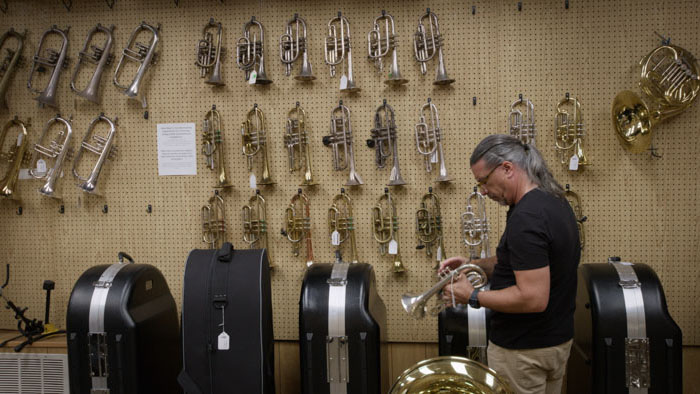 Jamie Egan inspects a trumpet among rows of brass instruments in Misfit Mentor.