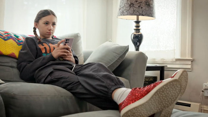 Bridget (Rylie Pennington) sits on a couch scrolling her phone in Not My Dog.