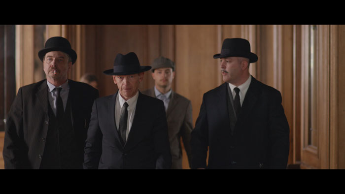 Three men in suits and hats walk through a wood-paneled hallway in The Gliwensbourg Chronicles.