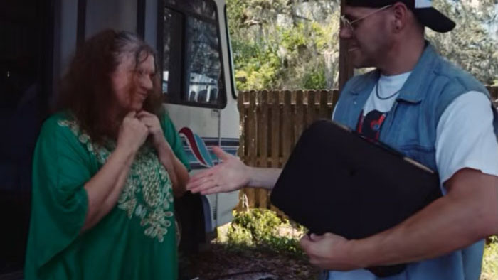 A man hands a black case to a woman outside an RV in a scene from The One Who Waits Below.
