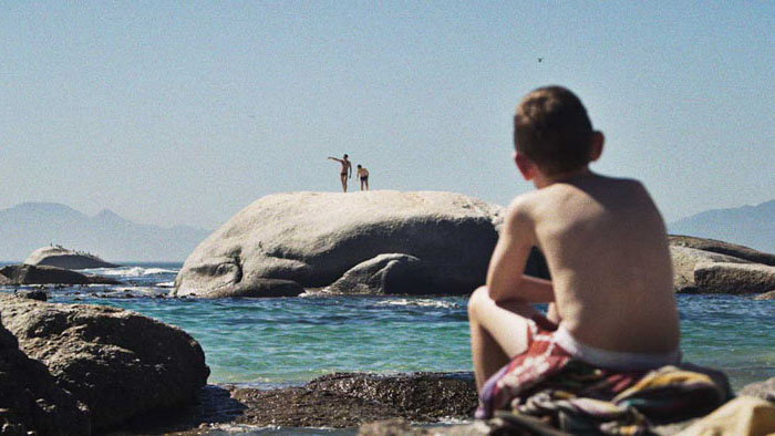 Marnus (Dawian van der Westhuizen) sits on rocks watching two distant figures on the shore in The Smell of Apples.